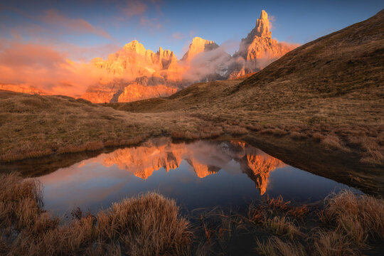 Pale di San Martino peaks and glowing orange clouds reflect in the Baita Segantini pond at sunset, Dolomites, Italy.