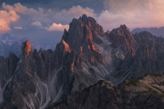 Rugged Dolomite peaks glow with warm sunset alpenglow under a dramatic pink and purple cloudy sky, Italy.