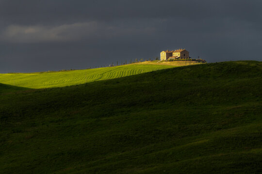 A lone stone farmhouse sits dramatically spotlit by a shaft of sunlight against a dark storm sky, green terraced fields sweeping down in the foreground. Val d'Orcia, Tuscany, Italy.