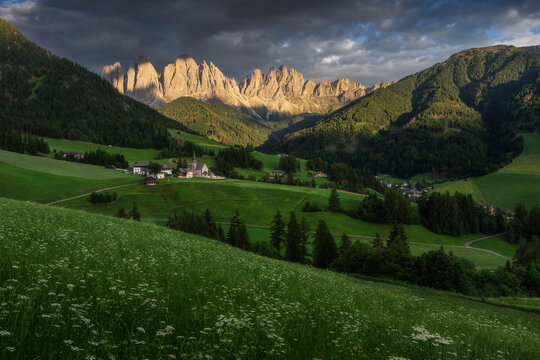 A picturesque Alpine village with Santa Magdalena church nestles in a green valley below sunlit Odle peaks, Val di Funes, Dolomites, Italy.