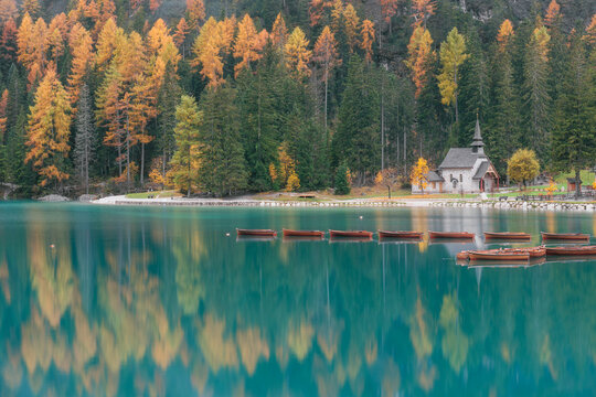 Wooden rowboats and a stone chapel reflect in the turquoise waters of Lago di Braies in autumn, Alto Adige, Italy.