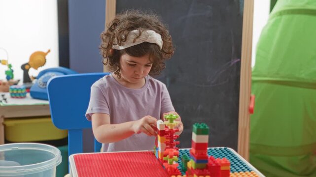 Girl builds colorful plastic blocks with hands, stacking gears and bricks at a play table inside a building; quiet concentration.