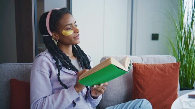 Person reads book on couch while wearing eye patches and relaxing at home during daytime