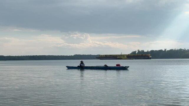 The silhouette of a fisherman angling while rowing across vast, calm waters beneath a cloudy sky. In the distance, yellow barges are moored in a peaceful bay, with the horizon framed by mangrove trees
