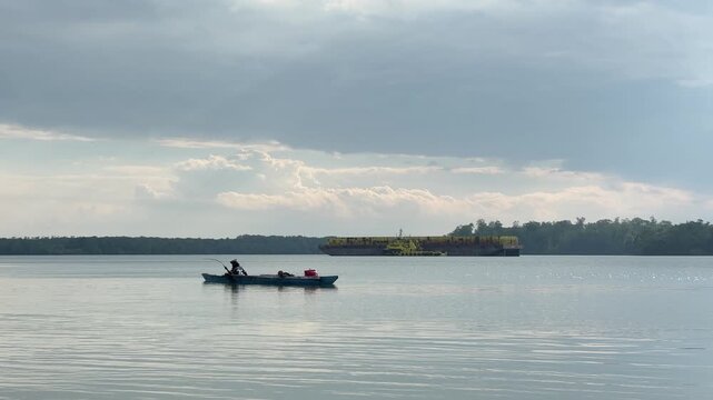 The silhouette of a fisherman angling while rowing across vast, calm waters beneath a cloudy sky. In the distance, yellow barges are moored in a peaceful bay, with the horizon framed by mangrove trees