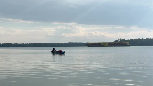 The silhouette of a fisherman angling while rowing across vast, calm waters beneath a cloudy sky. In the distance, yellow barges are moored in a peaceful bay, with the horizon framed by mangrove trees