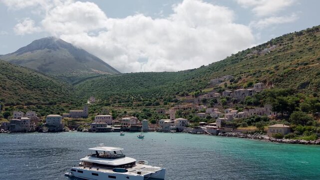 Aerial tracking shot of yachting in crystalline turquoise waters of Limeni village with old stone houses on shoreline with lush green mediterranean mountains in the backdrop under cloudy sky