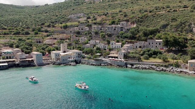 Drone shot of Limeni village turquoise waterfront ancient stone house towers surrounded by lush green mountains on a sunny day, fishing boats moored in turquoise sea water, Mani Peninsula, Peloponnese