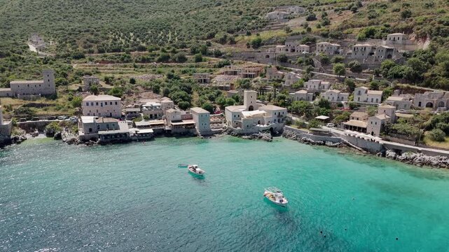 Aerial pullback shot of Limeni village on Mani peninsula, Turquoise waterfront old stone house towers on steep lush green mountains with mediterranean scrub, olive grove on a sunny day, Peloponnese
