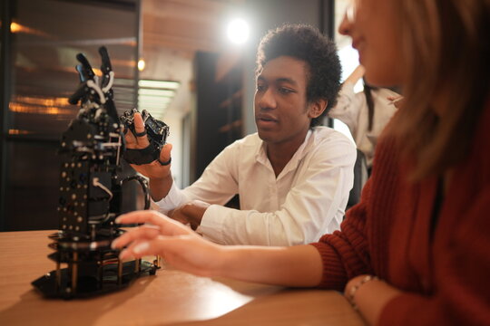 Student and classmate with a robotic hand discusses the robotic hand with a classmate close by in a bright modern training studio, celebrating diversity and teamwork in learning