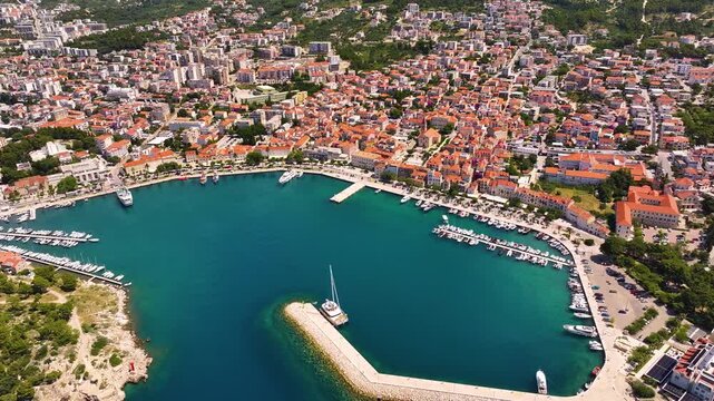 Makarska features a harbor with boats docked. Buildings surround the harbor. The town has residential and commercial areas. Roads lead to access points. The coastline is visible.