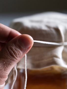 Brewing homemade scoby close up human thumb and index finger tying white string around linen cloth covering glass brewing jar in bright natural daylight