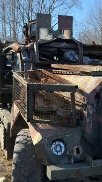 Front view of a ruined wheeled armored vehicle with rusted slat armor, shattered bulletproof glass, and a flat tire in a forest.
