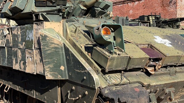 Close-up of a damaged military fighting vehicle hull showing a shattered headlight, explosive reactive armor blocks, and a white cross symbol.
