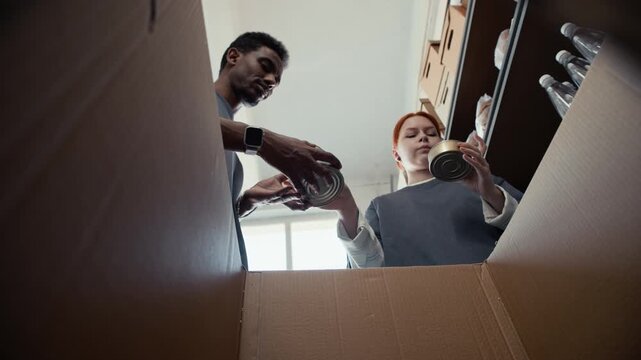 Young adult African American man and young adult Caucasian woman packing canned food into cardboard box, low angle view from inside box. Footage is perfect for humanitarian aid promos