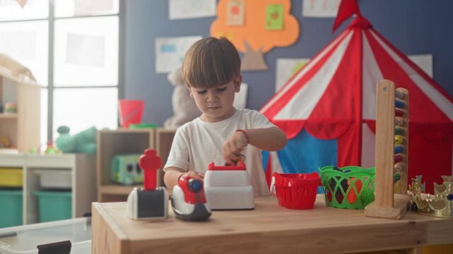 Boy presses toy cash register and taps buttons with bare hands in a building playroom filled with toys and a red tent; concentration play.