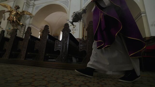 Catholic priest swinging a smoking thurible as he walks through an empty church, by rows of wooden pews and majestic religious artwork.