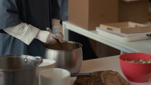 Volunteer wearing protective gloves stirring hot soup in metal pot during food distribution. Footage is ideal for humanitarian aid campaigns, charity promotions, and community service videos