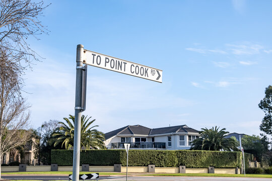A directional street sign reading "TO POINT COOK RD" at a suburban roadside. In the background, a large, contemporary double-story family home sits behind a manicured hedge in Melbourne Australia