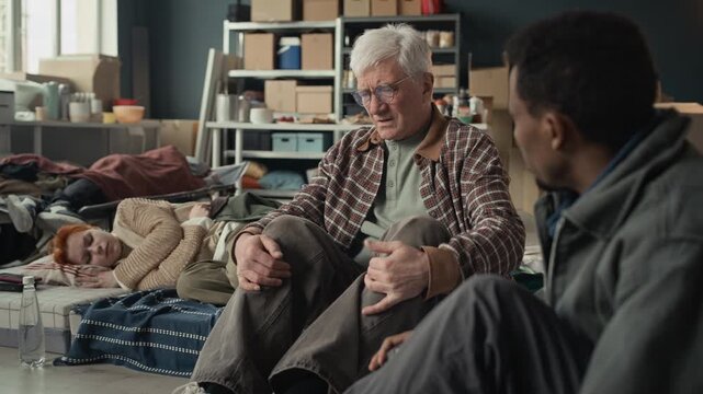 Caucasian senior man talking emotionally to Black young adult man in temporary shelter with young adult woman sleeping in background. Video perfect for humanitarian aid and social work campaigns