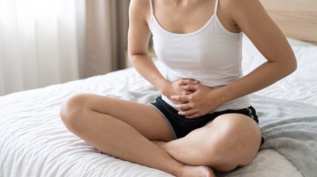 A young woman sitting on a white bed wearing a casual white tank top and black shorts while holding her stomach because she is experiencing sharp abdominal pain or cramps.