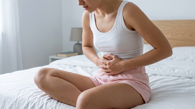 A young woman wearing a white tank top and pink shorts sits on a bed while holding her stomach in pain due to severe abdominal cramps and intense physical discomfort.