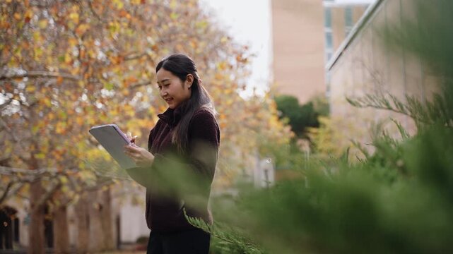 Smiling Asian woman drawing on a digital tablet with a pen while standing in an autumn park, Female student creative remote work, outdoor sketching, and freelance lifestyle