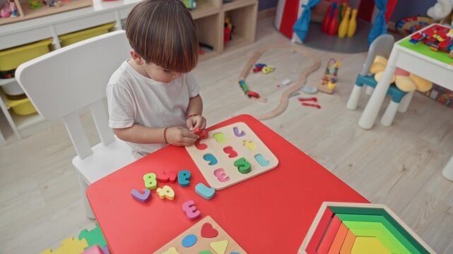 Boy placing a wooden alphabet puzzle piece on a red table with his right hand in a preschool playroom building; concentration learning.