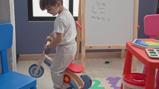 Boy straddling a wooden balance bike holds the handlebars and maneuvers between colorful chairs and a red activity table in a building playroom; playful exploration.