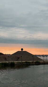 Vertical stock footage of a concrete step pyramid monument featuring a large ball on top by the river. Shot captures a vibrant orange sunset horizon, a distant bridge, and dramatic storm cloud