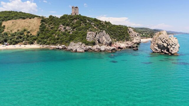 Drone flies low above the water in a circular motion around the coastal rocks, with Apollonia Tower visible above them on the hillside near Paralia Folias in Greece.