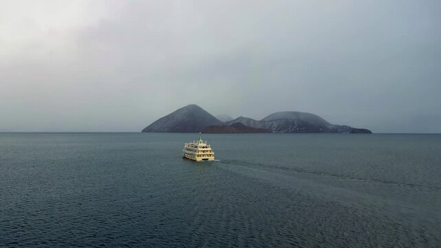 High-angle aerial view of a sightseeing tour ferry sailing through the calm waters of Lake Toya with volcanic islands in the background, winter in Japan.