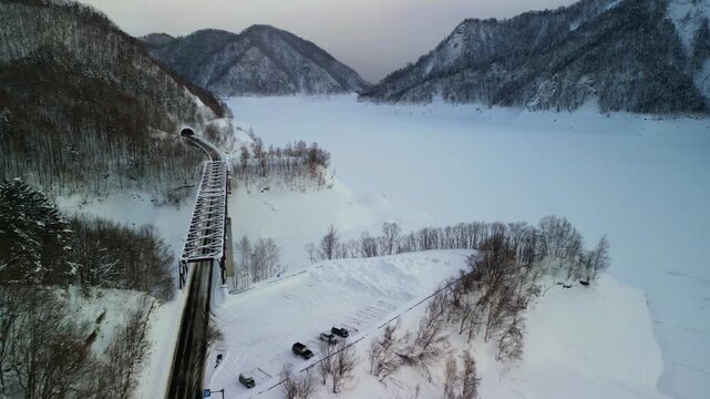 Scenic drone shot of a steel truss train bridge leading into a mountain tunnel alongside a vast frozen lake covered in snow.