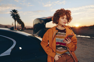 Naklejka premium Woman lifestyle on road at sunset near car, curly hair, orange jacket, holding tablet, calm smile, evening light, travel portrait, parked vehicle background