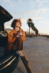 Naklejka premium Woman lifestyle outdoors near car at sunset, curly hair smiling and holding coffee, casual jacket, relaxed street mood, soft breeze, warm golden light, palm trees blurred background