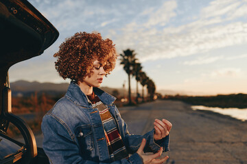 Naklejka premium Woman lifestyle outdoors sunset, curly hair denim jacket scarf by car, evening road walk, relaxed posture, warm golden light, coastal scenery, natural colors, soft breeze