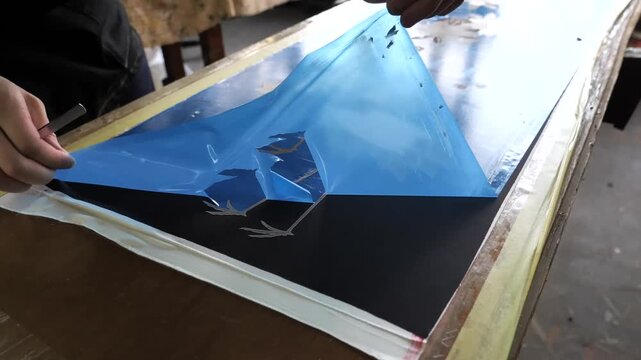 Artisan peels a blue plastic stencil film revealing a painted crane and red flower design beneath