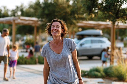 A woman is joyfully smiling in a vibrant outdoor park, surrounded by greenery and families, capturing a moment of happiness and community in a lively atmosphere.