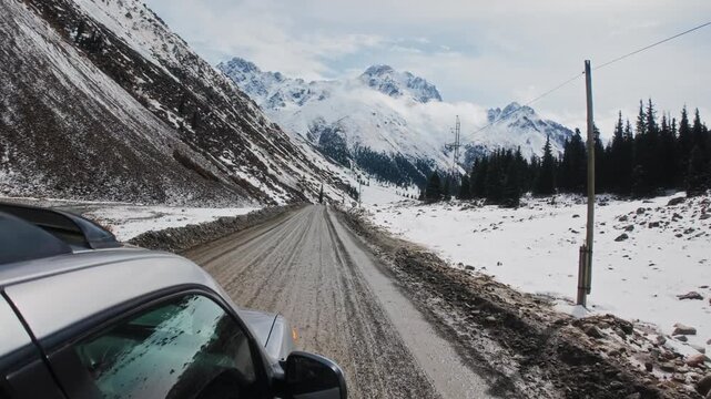 Car Driving on Dirt Mountain Road in Snowy Winter Landscape with Rocky Mountain Peaks