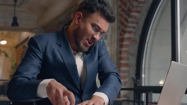 Overworked multitasking businessman Indian business man many task talking phone writing noting in notebook using laptop Arabian male guy panick manager with smartphone documents anxious in office