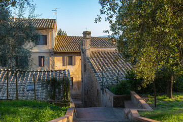 Obraz premium Stone Tuscan house with a tiled roof and garden path, surrounded by trees and greenery in warm evening light. A quiet countryside scene near Montaione, Italy.