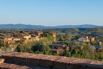 Panoramic view of a San Gimignano Tuscan village with rooftops, fields, and distant mountains under a clear blue sky. A classic rural Italian landscape. © Lukasz