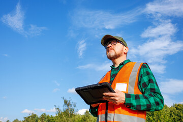 Fototapeta premium Climatologist or environmental supervisor in protective uniform analysing weather data on a digital tablet in front of a green forest and blue sky.