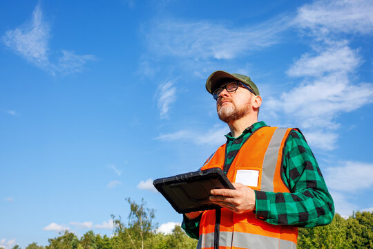 Climatologist or environmental supervisor in protective uniform analysing weather data on a digital tablet in front of a green forest and blue sky.