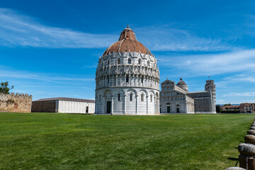 Fototapeta premium PISA ITALY APRIL 5 2026: Pisa Baptistery and the Leaning Tower of Pisa viewed from the grass under a bright blue sky. A famous Italian landmark in a classic wide-angle composition.