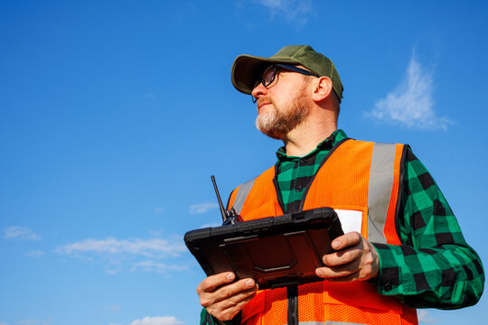 Environmental engineer or field supervisor conducting land monitoring and data collection using rugged digital tablet under blue sky. Male forestry inspector or ecologist.