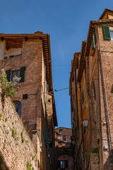 Fototapeta premium Narrow medieval street in Siena with tall brick buildings and a bright blue sky overhead. The composition emphasizes the vertical lines and old-world atmosphere.