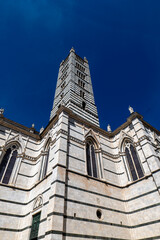 Fototapeta premium Low-angle view of Siena Cathedral and its tall bell tower against a deep blue sky. The image emphasizes the monument’s dramatic height and white marble details.