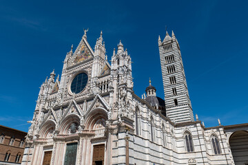 Fototapeta premium Wide view of Siena Cathedral with its ornate facade and bell tower rising above the square. The bright blue sky creates a striking contrast with the white marble architecture.
