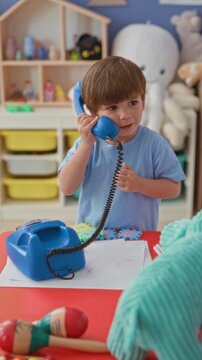 Boy holding blue toy telephone to ear at a preschool building playroom table, hand on receiver and drawing paper nearby; curiosity imagination.